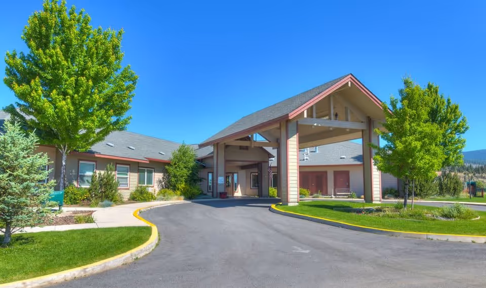 Exterior view of Eagle Lake Village facility showing a covered entrance with a peaked roof, surrounded by green trees and landscaped grass areas under a clear blue sky.
