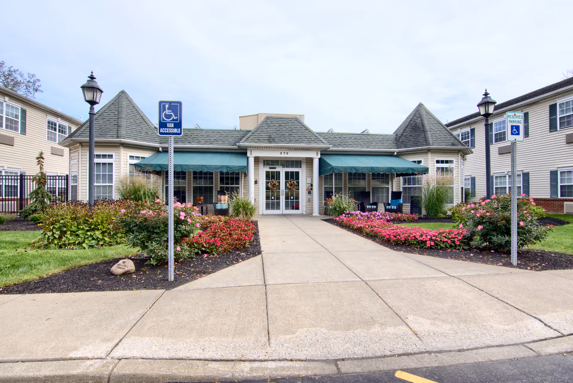 Front exterior view of Peregrine Senior Living at Cheektowaga building with a paved walkway leading to double glass doors. The building has a green awning over the entrance and windows, landscaped flower beds with pink and red flowers on either side of the walkway, and two lamp posts with handicap parking signs.