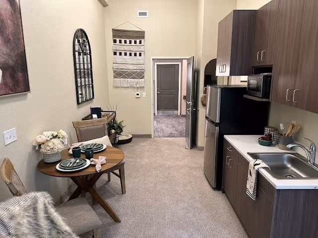 Interior view of a senior living facility kitchen and dining area with a small round wooden table set for two, two chairs, a stainless steel refrigerator, microwave, sink, and wooden cabinets. The walls are beige with decorative wall hangings and a doorway leading to another room.