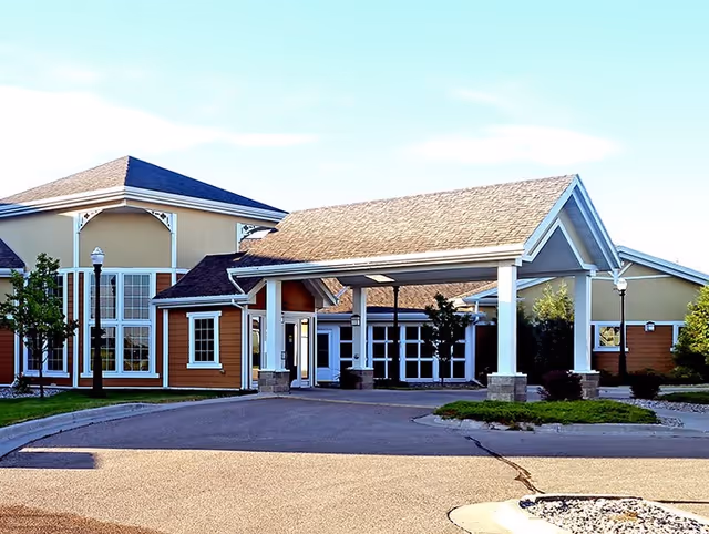 Exterior view of Good Samaritan Society - Fargo building with a covered entrance, large windows, and a driveway in front. The building has a beige and brown color scheme with a shingled roof and some landscaping including trees and bushes.