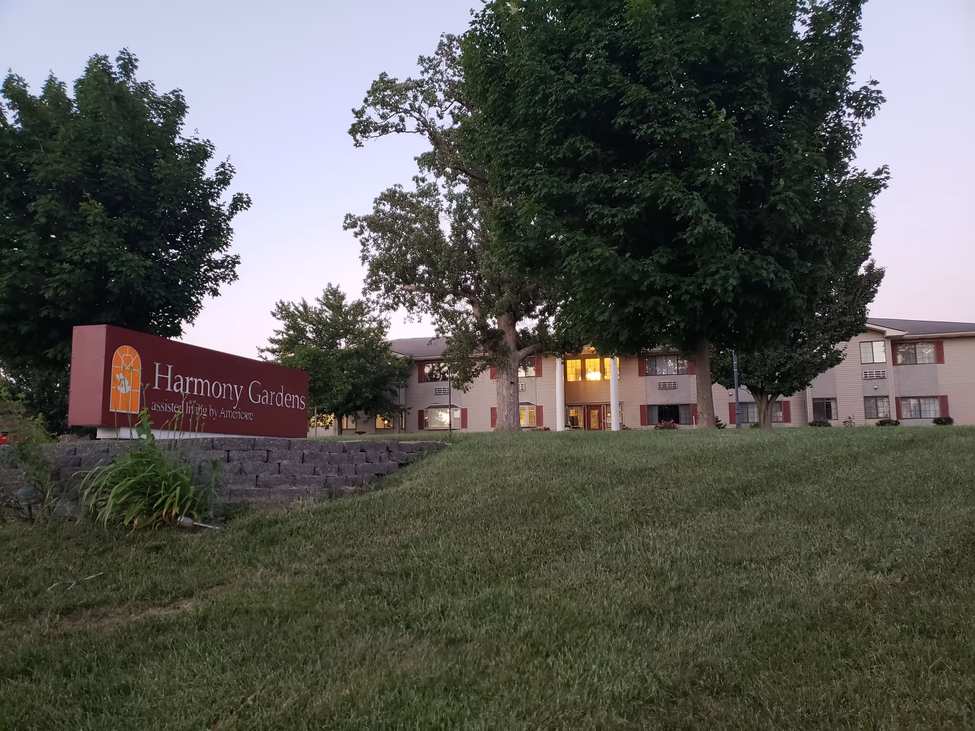 Exterior view of Harmony Gardens facility with a red sign and large trees in front.