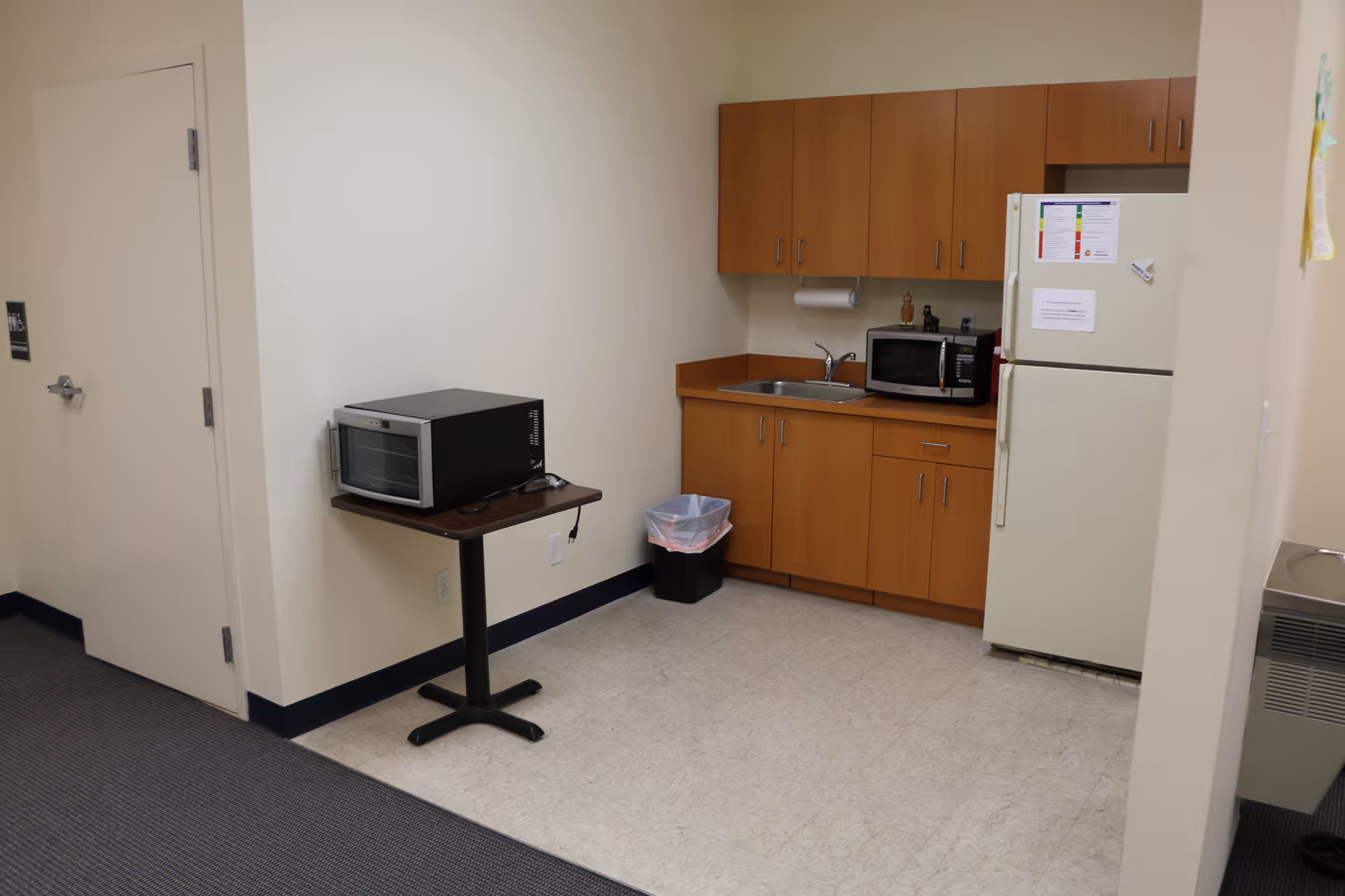 Compact kitchenette with wooden cabinets, sink, microwave, and a full-sized refrigerator.
