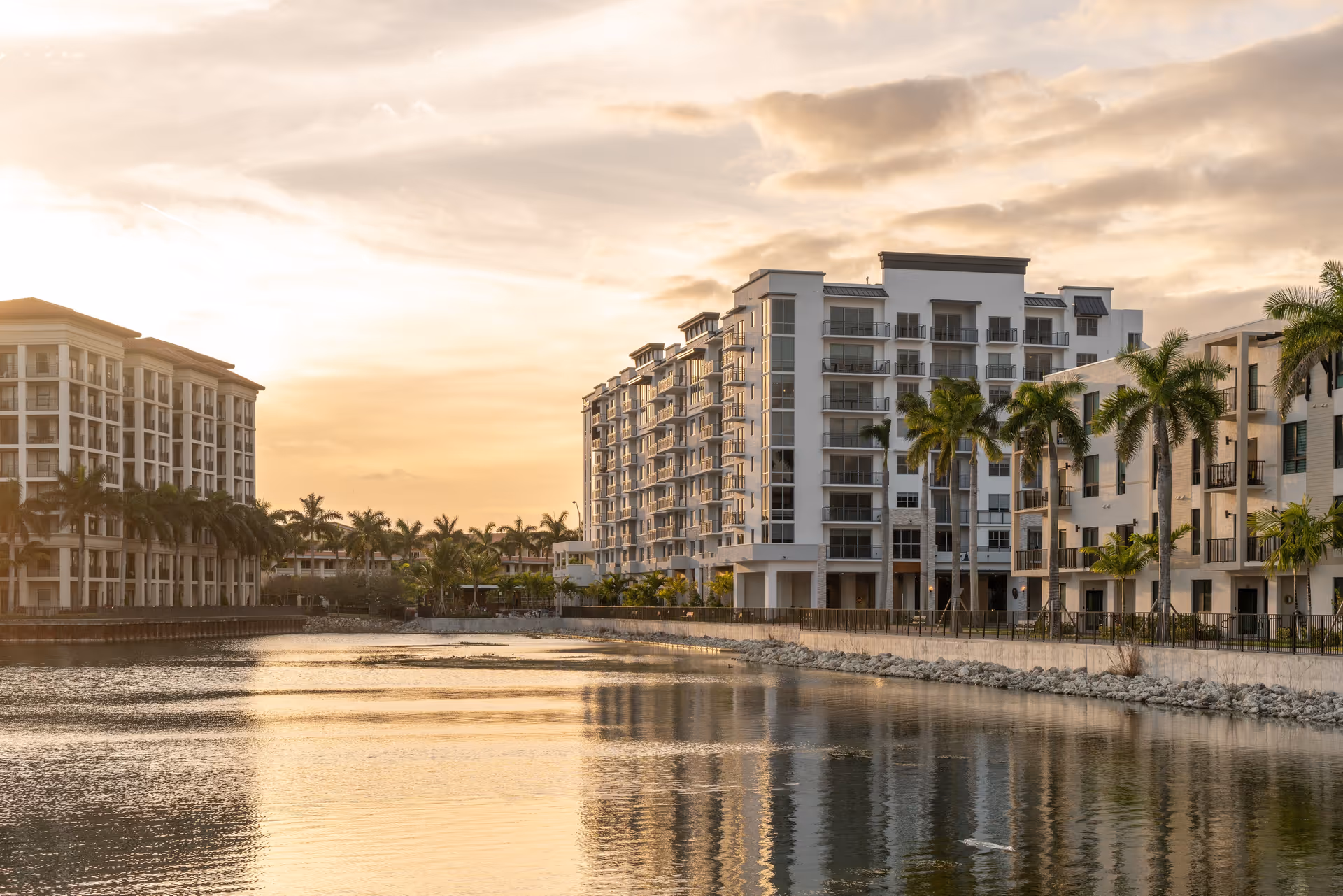 Sunset view of modern waterfront residential buildings with palm trees reflected in the water.