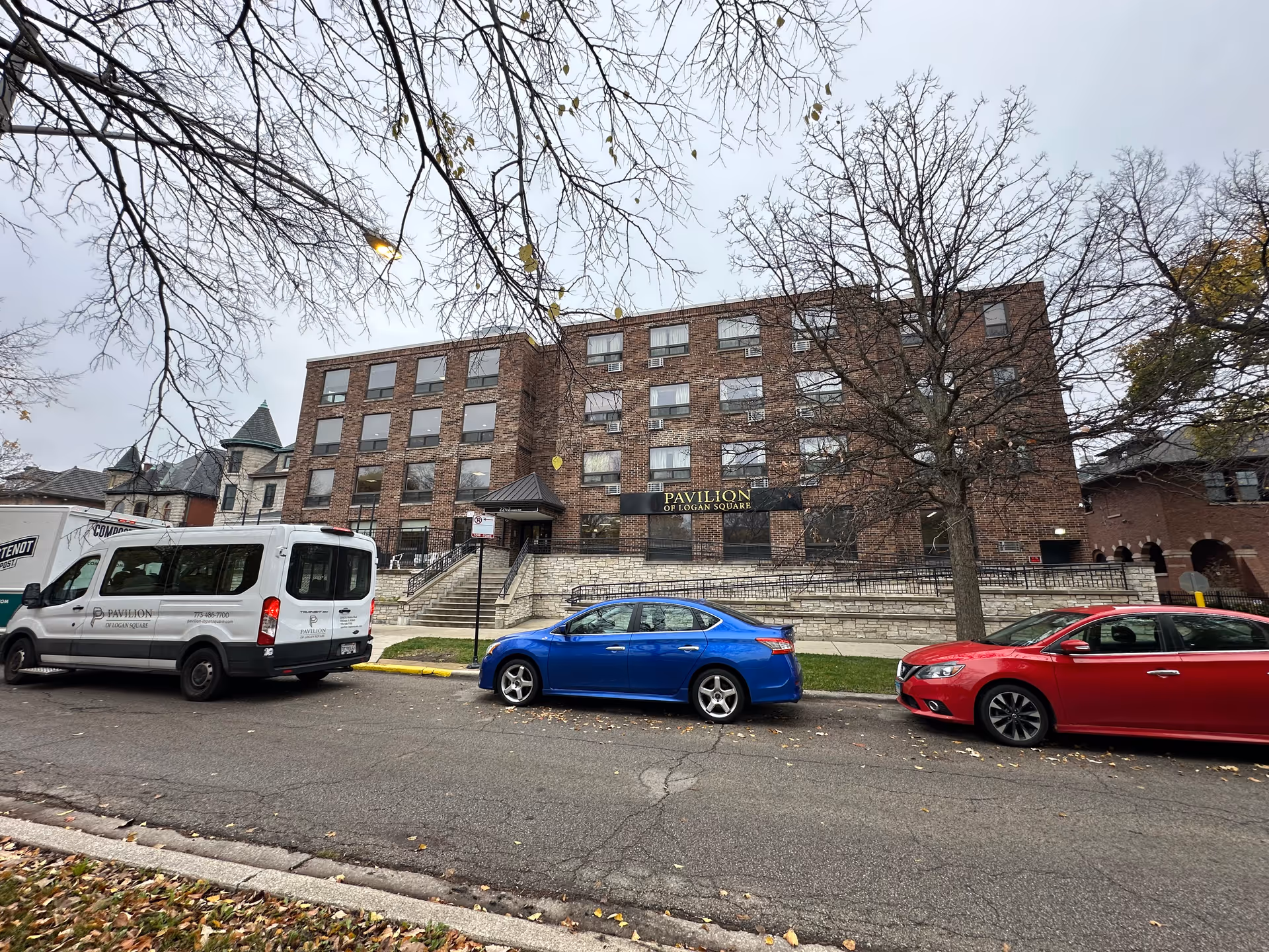 Front view of the brick Pavilion of Logan Square senior living building with parked cars along the street.
