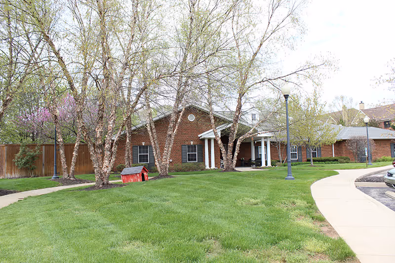 Brick senior living facility with front entrance, green lawn, trees, and street lamps.