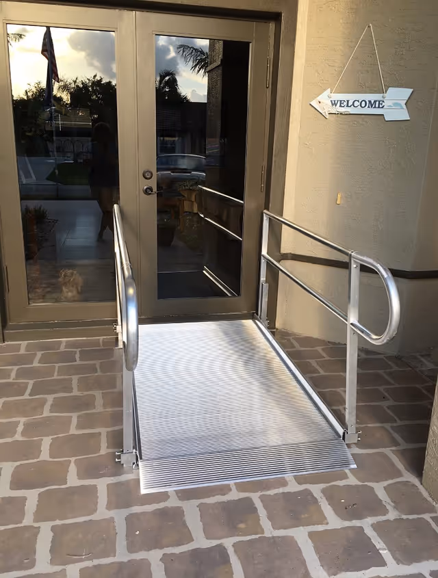 Metal ramp with handrails leads to glass double doors at facility entrance.