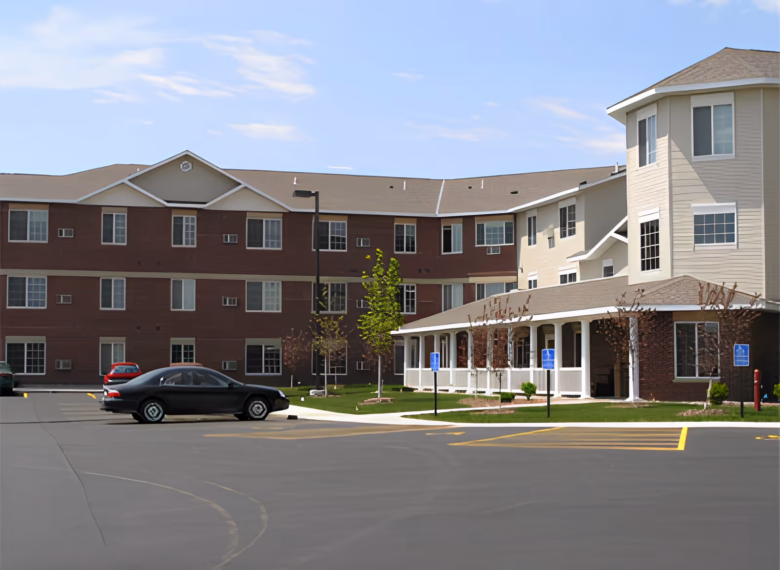 Front exterior of a three-story senior living building with a covered entrance, small lawn, and parked cars in the lot.