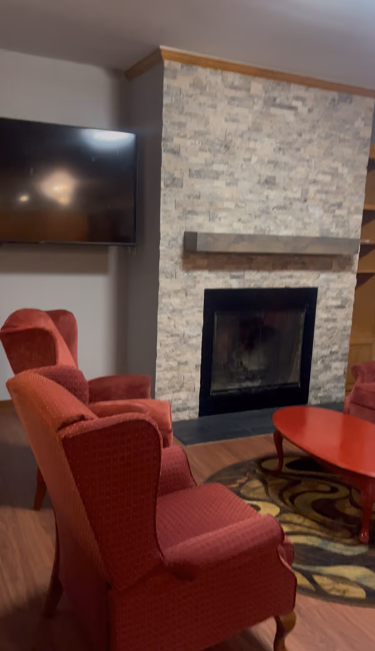 Cozy living room with red upholstered armchairs, a stone fireplace, and a wall-mounted TV.