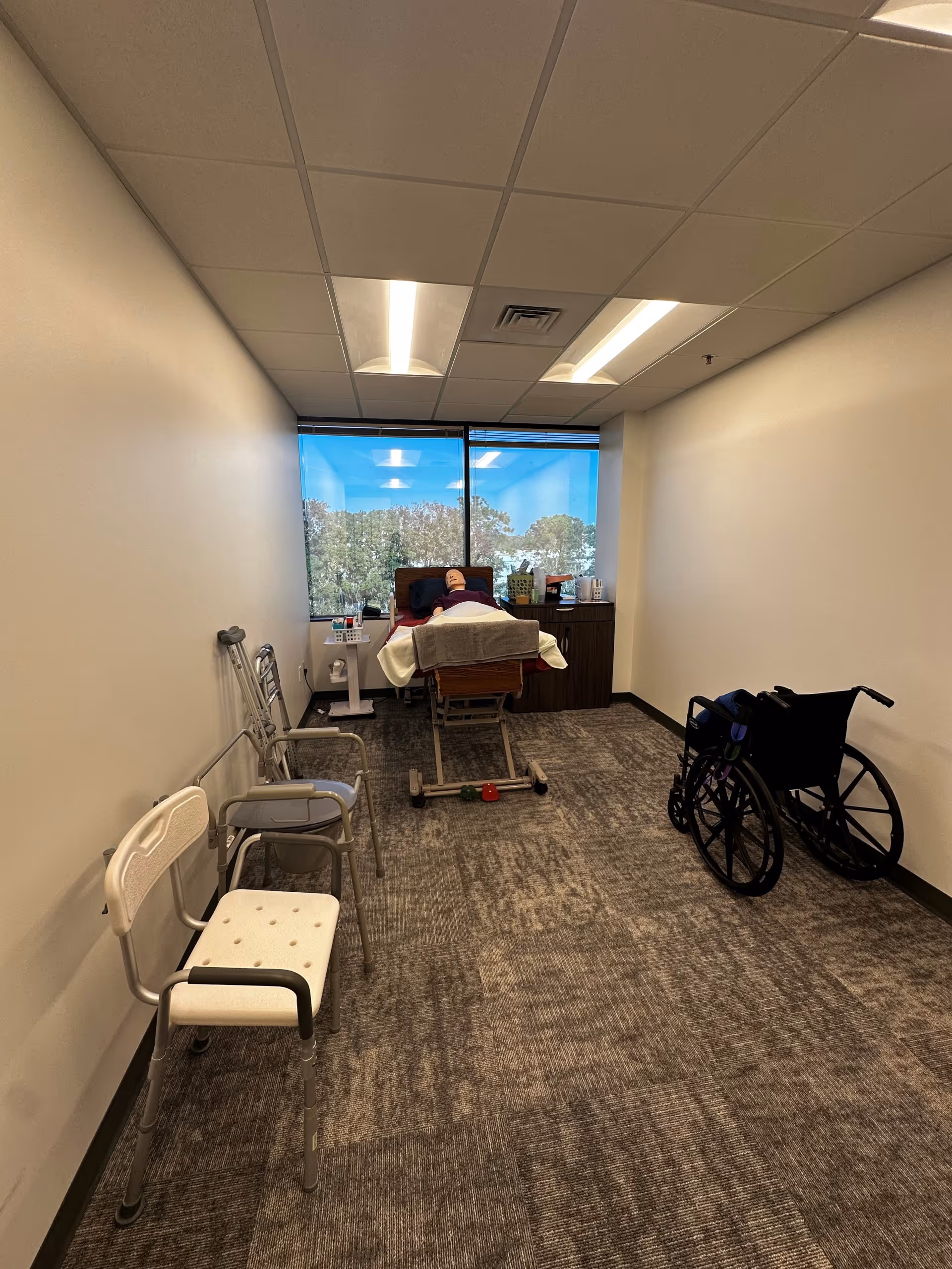 Narrow treatment room with a hospital bed, wheelchair, and several chairs near a windowed back wall.