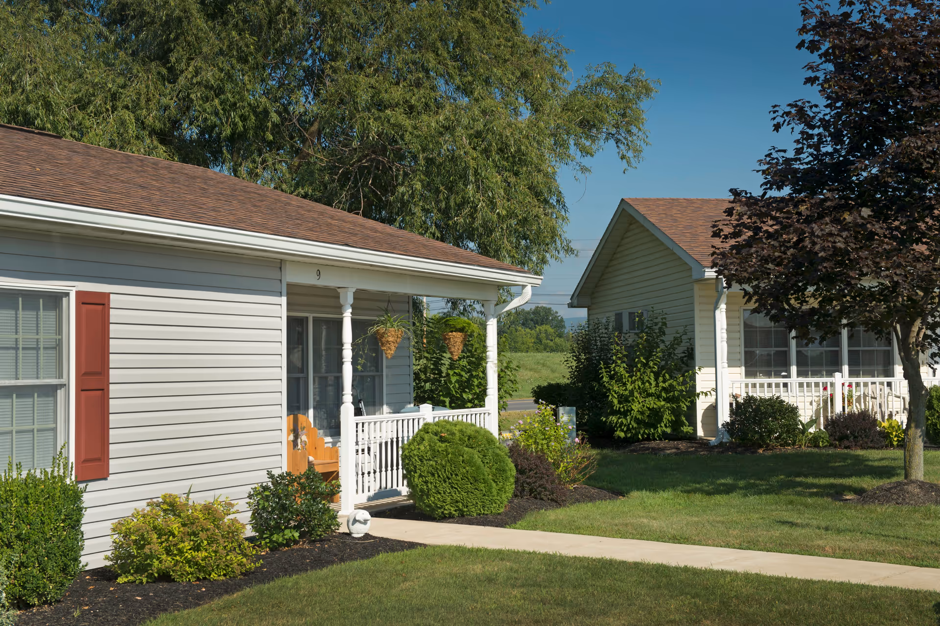 Single-story cottage-style units with front porches, hanging planters, and neatly maintained lawns under a clear sky.