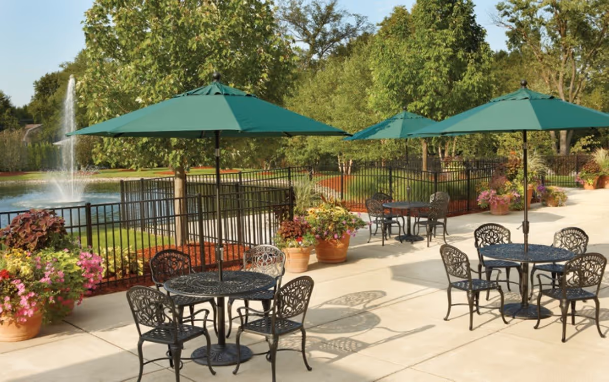 Outdoor patio with metal tables and green umbrellas overlooking a pond and fountain, surrounded by potted flowers and trees.
