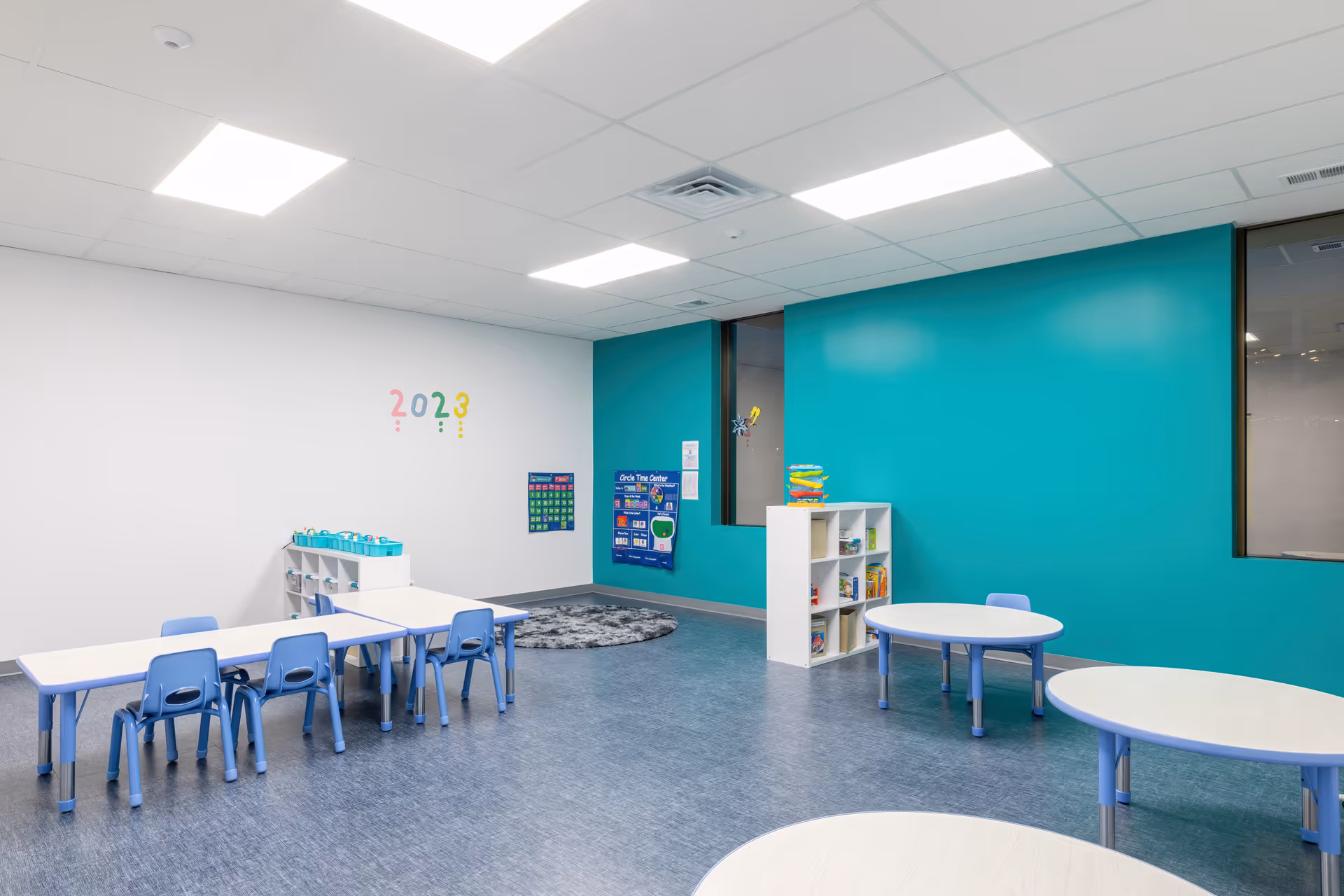 Bright classroom space with kid-sized tables, blue chairs, teal wall, and shelves.