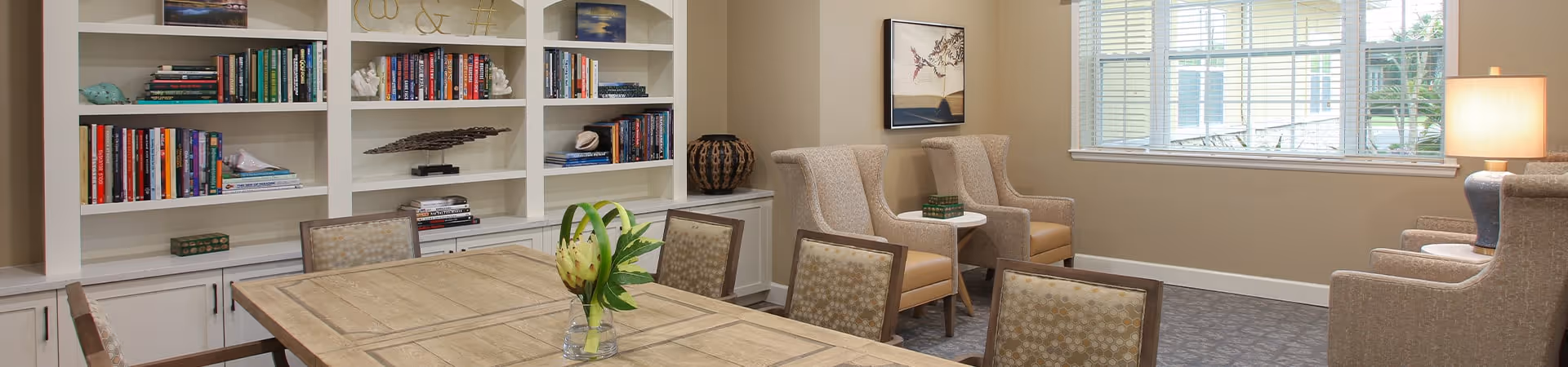 A cozy interior room with a wooden table surrounded by chairs, a built-in bookshelf filled with books and decorative items, two beige armchairs with a small side table between them, a window with blinds letting in natural light, and a table lamp on a side table.