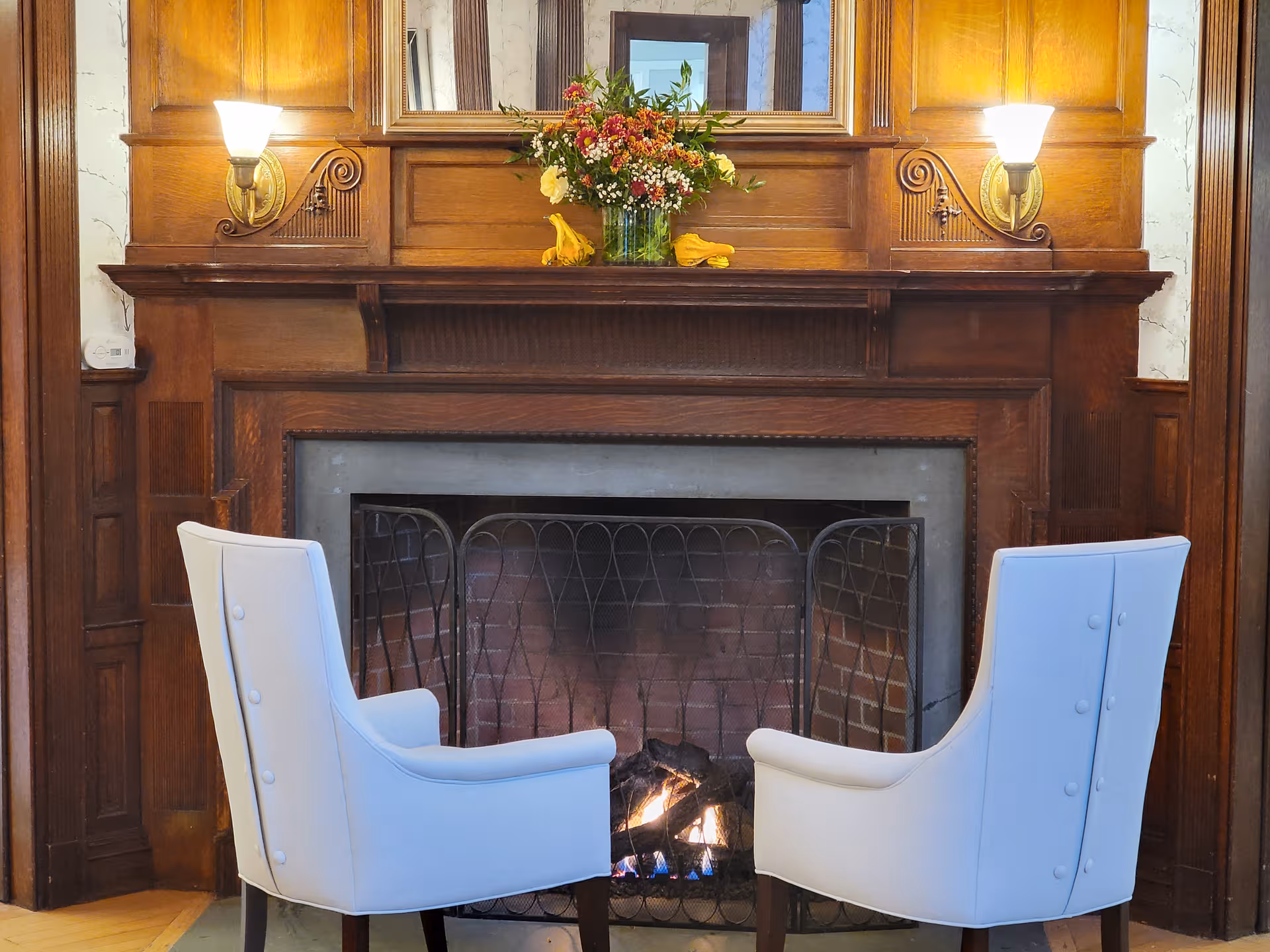 Cozy living room with a wooden fireplace, two light-blue chairs, and floral mantel decor.