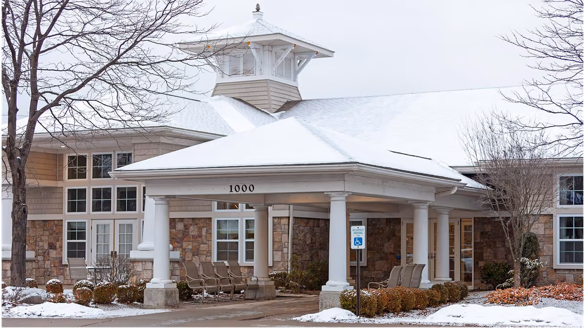 Covered portico entrance with white columns and the number '1000' on the pediment, shrubs and a light dusting of snow in front of a senior living building.