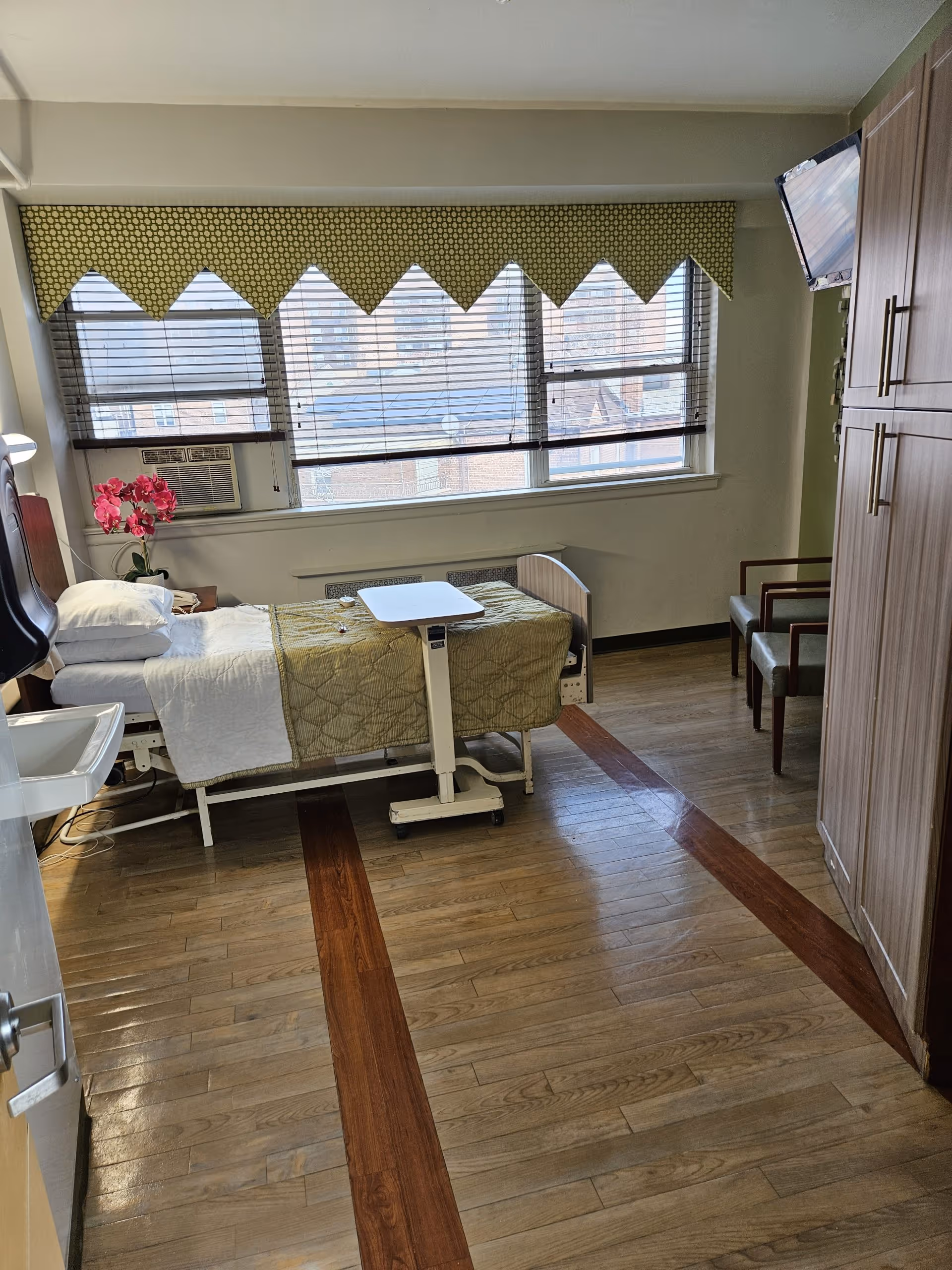 A senior care facility room with a hospital-style bed covered with a green quilt and white sheets, a small overbed table, two chairs, a wall-mounted television, a large window with blinds and a patterned valance, a wooden floor with darker wood inlays, and a cabinet on the right side.