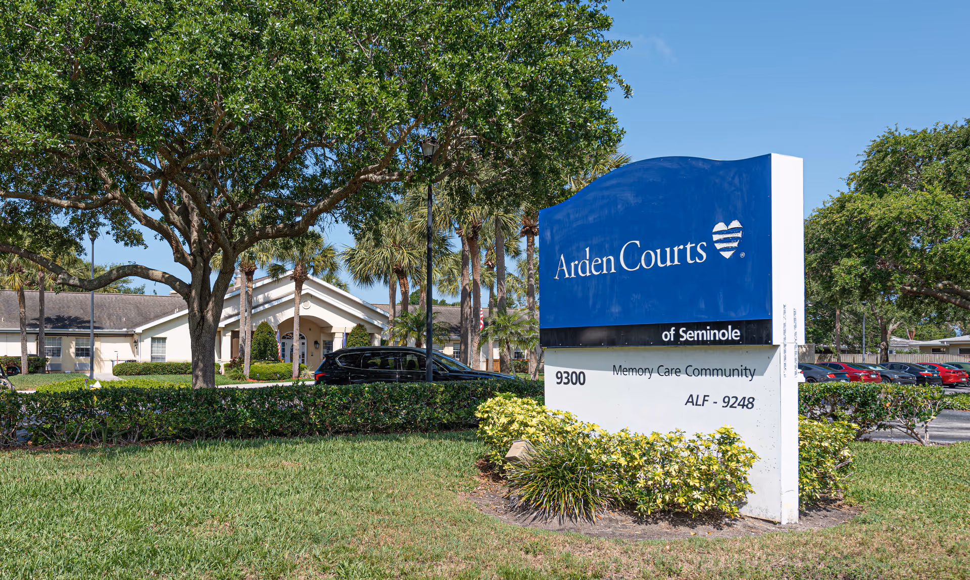 Outdoor view of Arden Courts - ProMedica Memory Care Community in Seminole, showing a large blue and white sign with the facility name and address, surrounded by green grass, bushes, and trees with a building and parked cars in the background under a clear blue sky.
