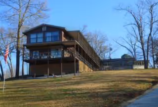 Two-story wooden lodge-style building on a grassy hill with a deck and stairs.