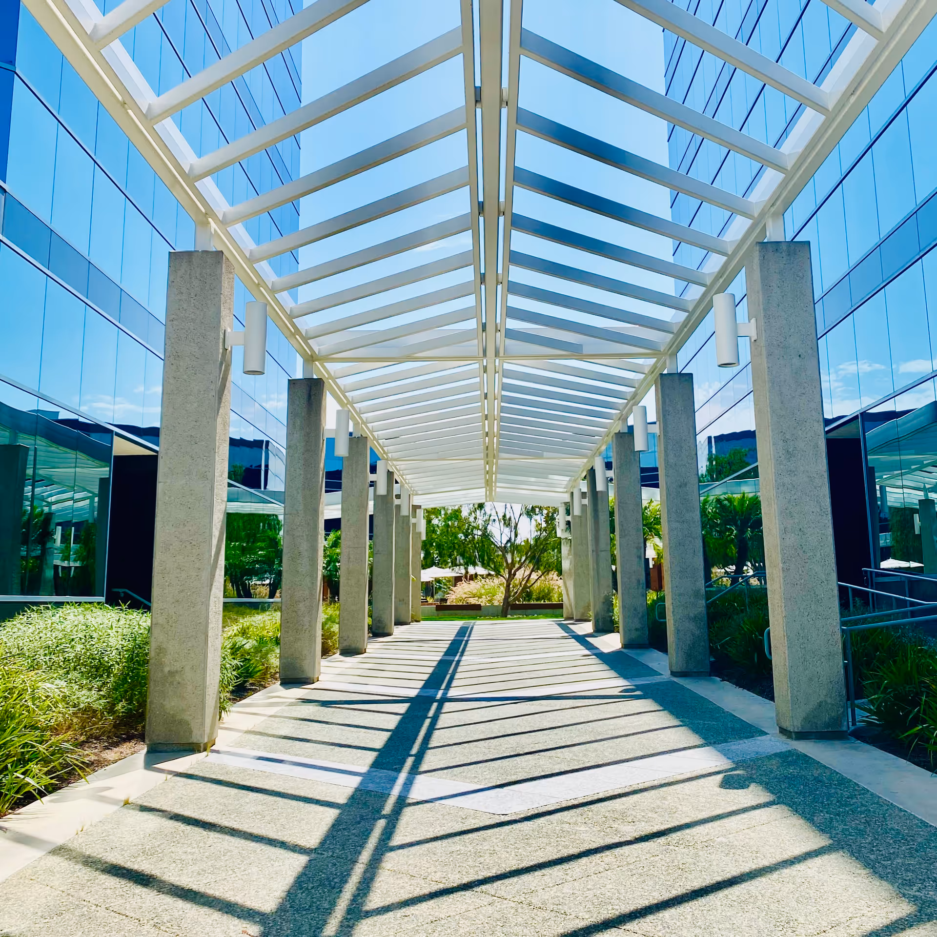 Covered outdoor walkway with glass walls, concrete pillars, and striking striped shadows.