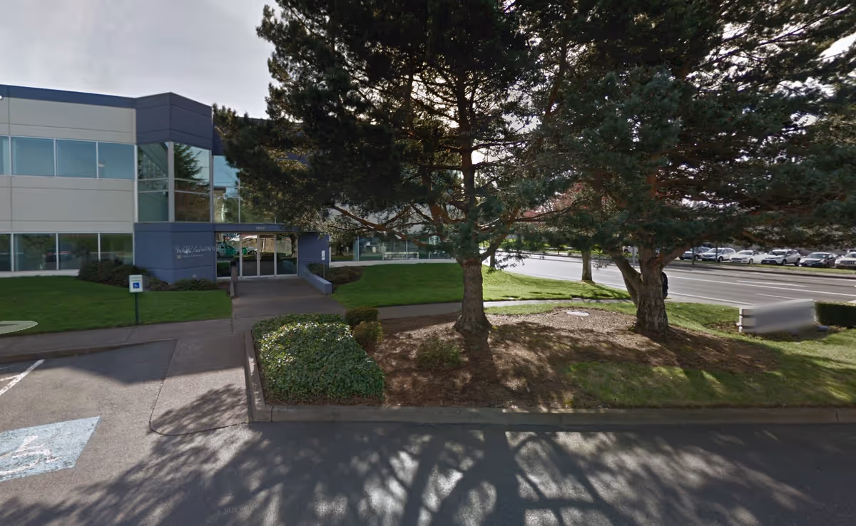 Modern facility exterior with glass entrance, trees, and a parking lot.