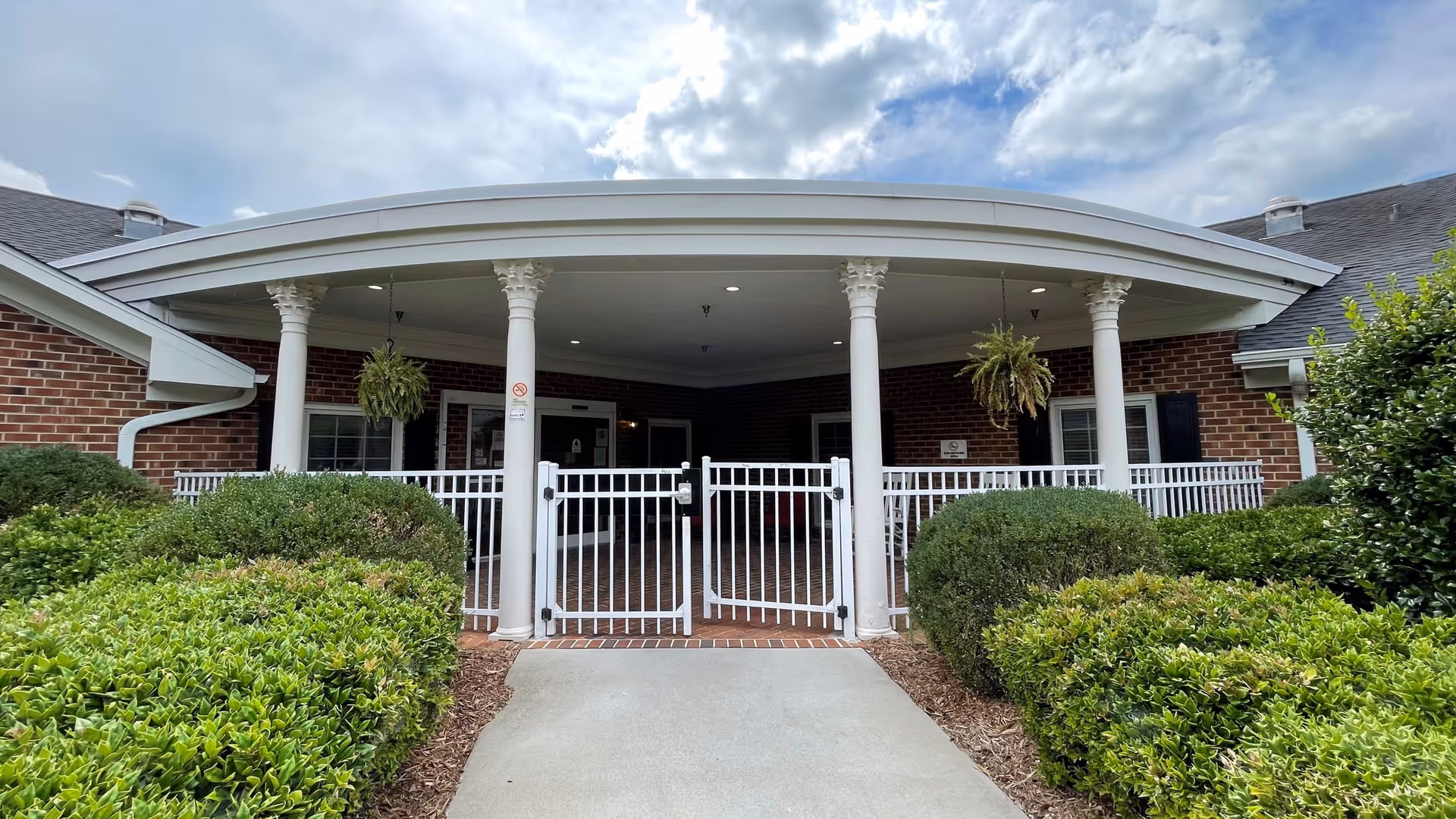 Brick senior living facility exterior with curved portico, white columns, gate, and trimmed hedges.