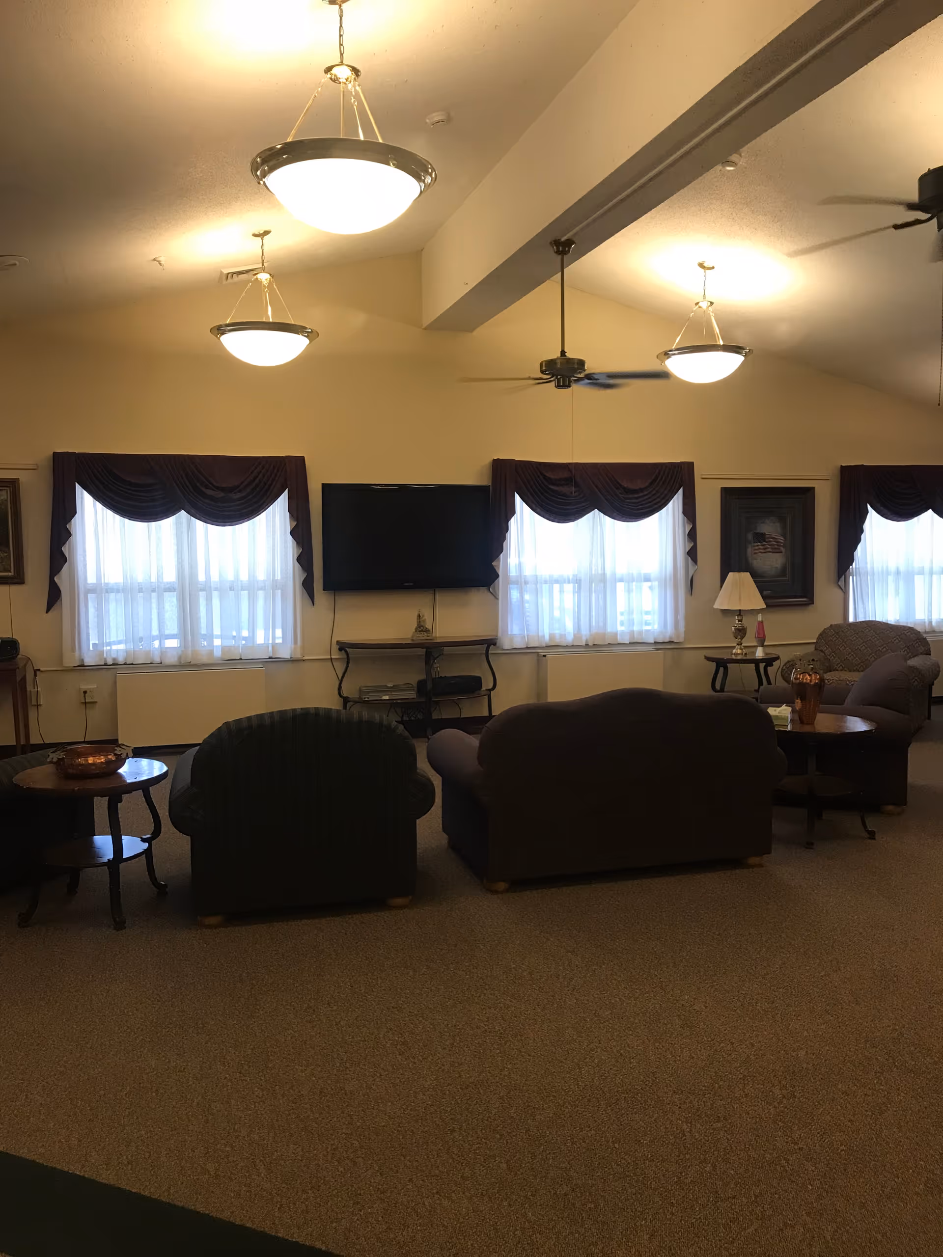 A cozy living room area with three armchairs and a sofa arranged around a wall-mounted flat screen TV. The room has three windows with sheer white curtains and dark valances. There are two side tables with lamps and decorative items, and ceiling lights and fans are visible on the ceiling.