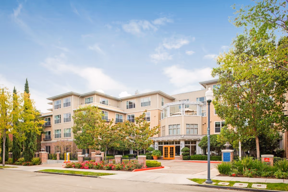 Four-story assisted living facility with a landscaped entrance, trees, and a circular driveway under a blue sky.
