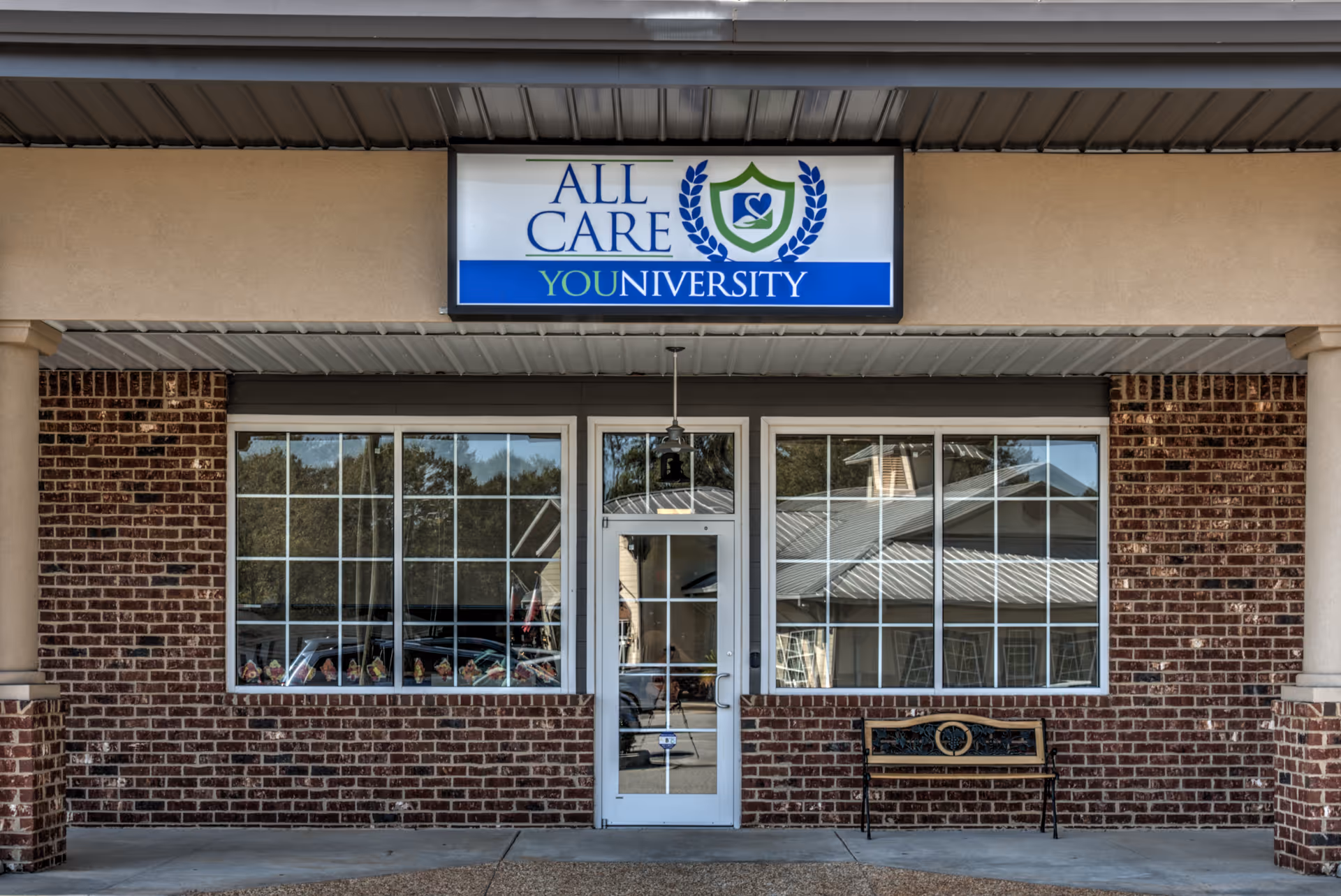 Brick storefront with 'All Care' sign, large windows, glass door, and bench outside.