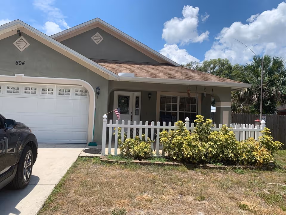 Front exterior of a single-story house with a garage, white picket fence, porch and shrubs under a blue sky.