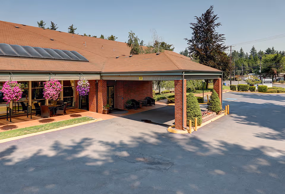 Exterior view of Firwood Gardens facility entrance with a covered drop-off area supported by brick columns, hanging flower baskets, outdoor seating, and surrounding greenery under a clear sky.