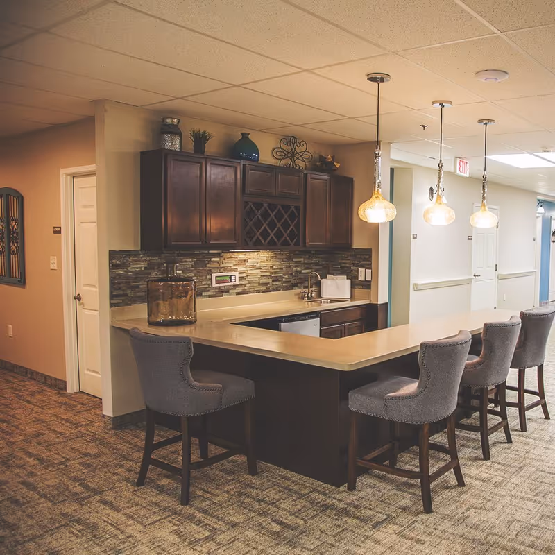 Interior view of a senior living facility kitchen area with a long countertop bar and four upholstered bar stools. The kitchen features dark wooden cabinets, a tiled backsplash, a sink, and decorative items on top of the cabinets. Three pendant lights hang above the countertop, and a hallway with doors is visible in the background.