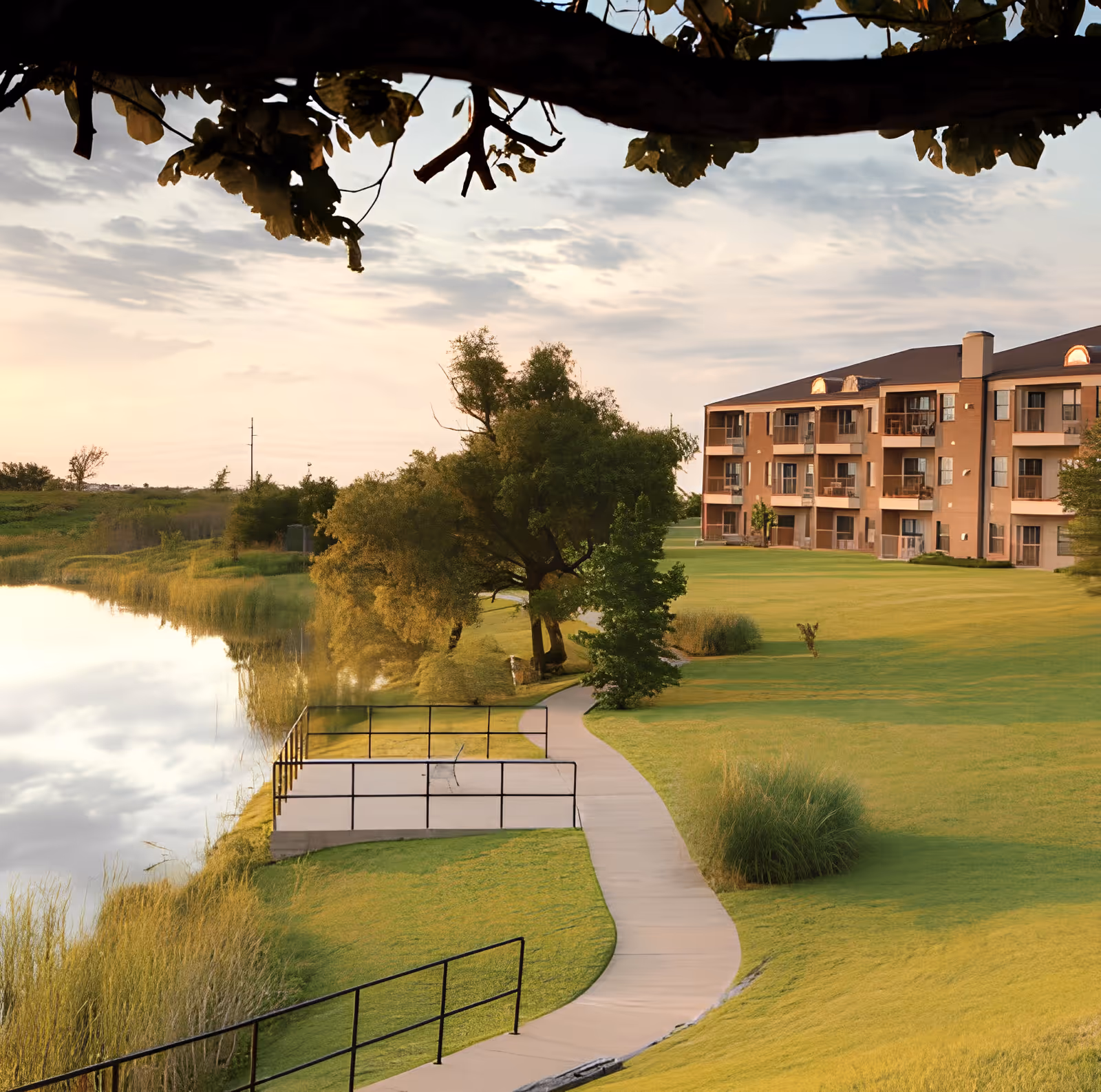 Lakeside view of a multi-story senior living building with a winding walkway, grassy lawn, trees, and a reflective pond.