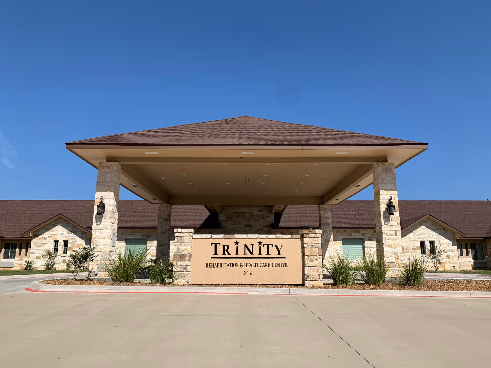 Front exterior view of Trinity Rehabilitation & Healthcare Center building with a covered entrance supported by stone pillars, clear blue sky in the background.