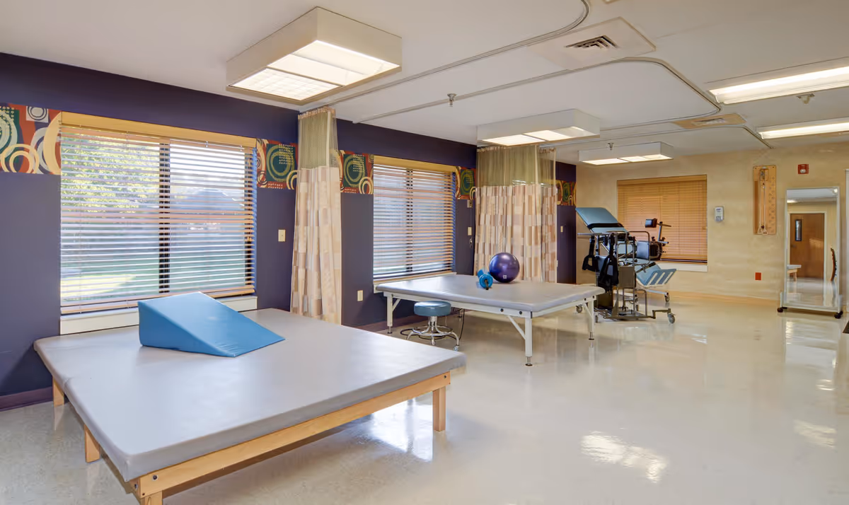 Bright therapy room with two treatment tables, exercise equipment, and large windows.