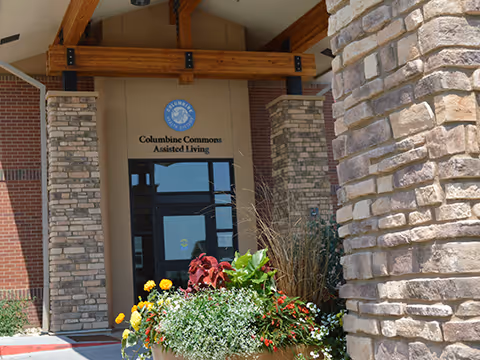 Entrance of Columbine Commons Assisted Living facility showing a covered entryway with stone pillars, wooden beams, and a large planter filled with colorful flowers in front.