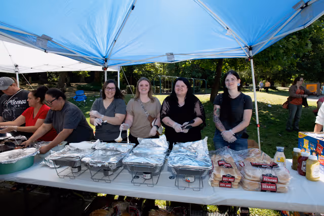 Group of people serving food under a blue canopy outdoors.