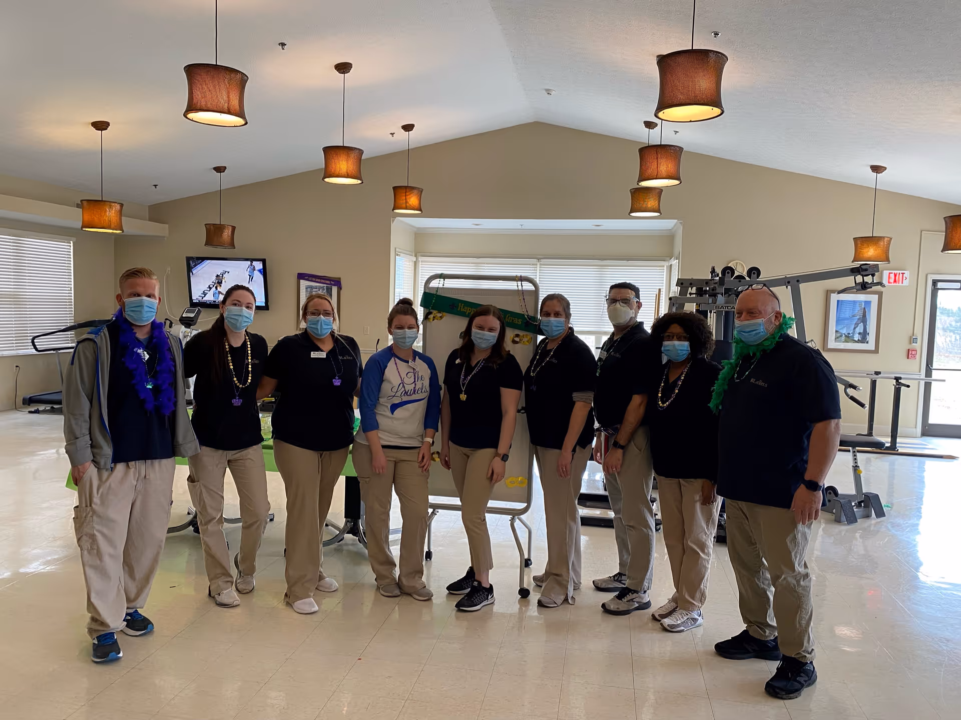 A group of nine staff members wearing masks and casual work attire standing together in a spacious room with exercise equipment and pendant lights hanging from the ceiling. They are posing for a photo and some are wearing festive beads and accessories.