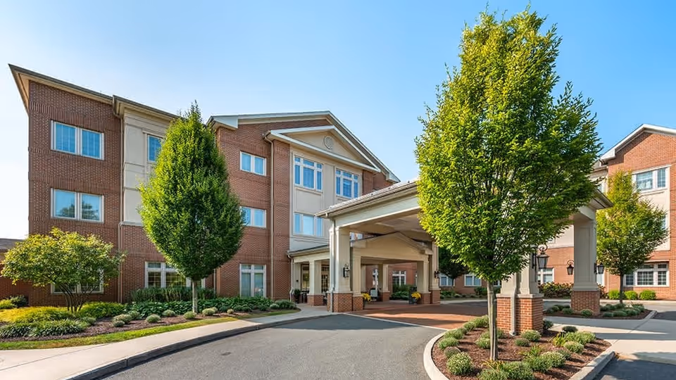 Exterior view of a multi-story senior living facility building with a covered entrance driveway, surrounded by landscaped greenery and trees under a clear blue sky.