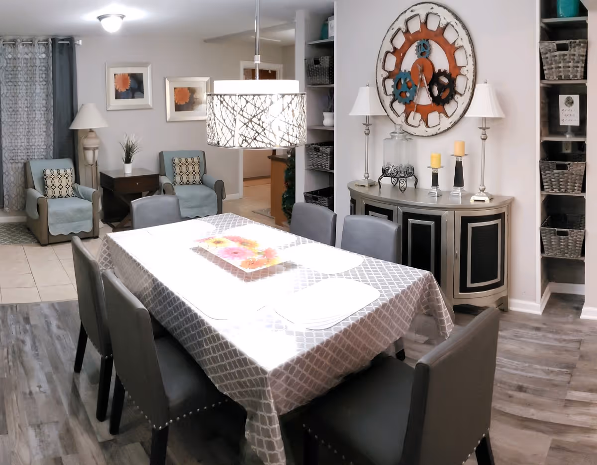 A dining area with a rectangular table covered with a patterned tablecloth and six gray chairs around it. Above the table hangs a modern light fixture with a decorative shade. In the background, there are two armchairs with cushions and a small side table with a lamp and a plant. On the right wall, there is a large decorative clock above a sideboard with candles and lamps. Shelves with baskets are visible on the far right.