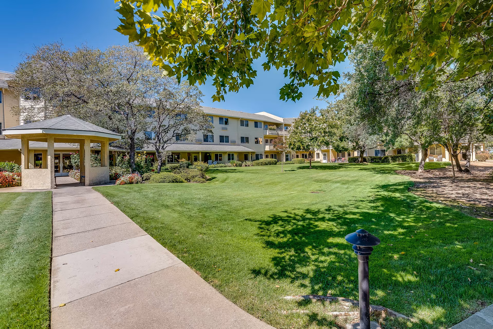 A paved walkway leads across a manicured lawn to a covered entrance of a multi-story senior living building surrounded by trees.