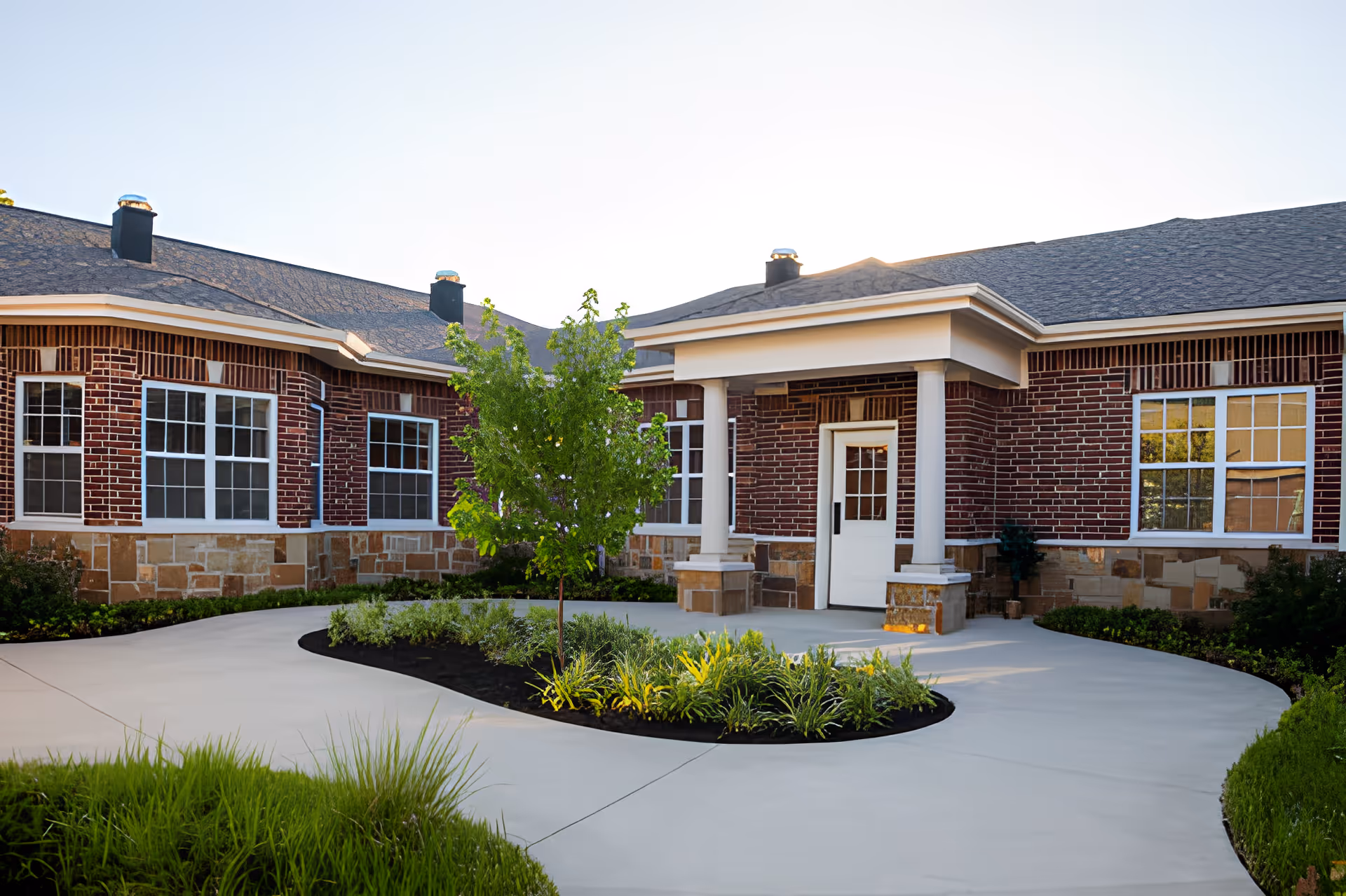 Front entrance of a single-story brick memory care building with a curved concrete walkway and landscaped planting beds.