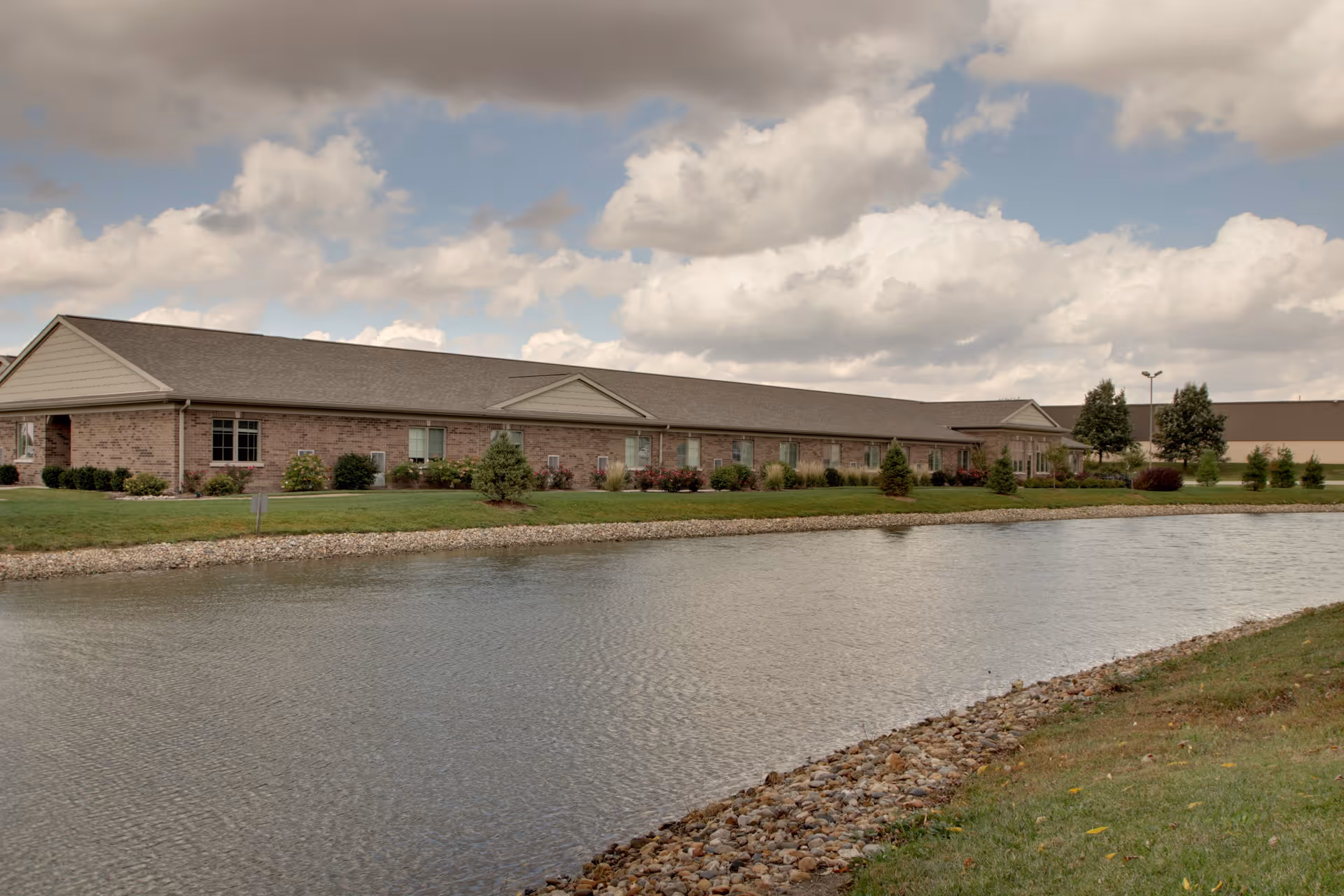 A long, single-story brick building with a gabled roof situated behind a pond with a rocky edge. The building is surrounded by well-maintained grass, shrubs, and small trees under a partly cloudy sky.