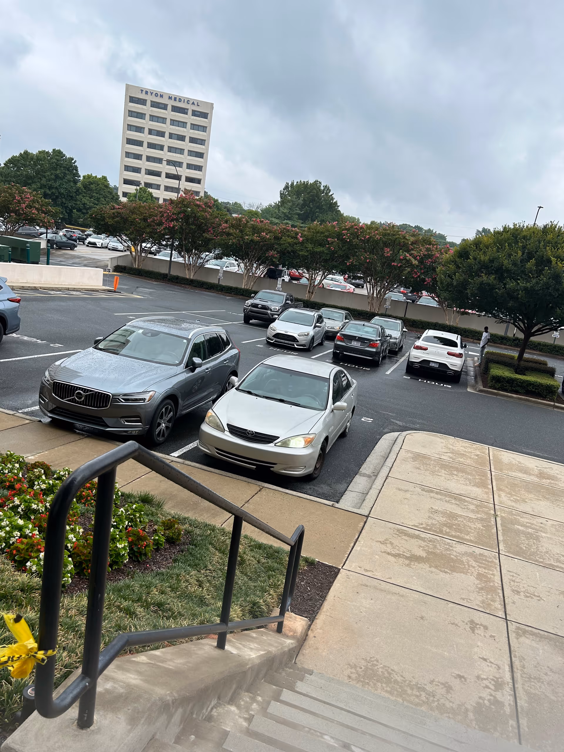 Exterior parking area with multiple cars and a distant building, railing and steps in foreground.