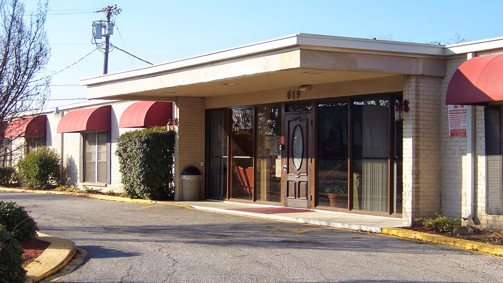 Street-level view of the front entrance of a single-story senior living building with glass doors, a covered entry, and red awnings.