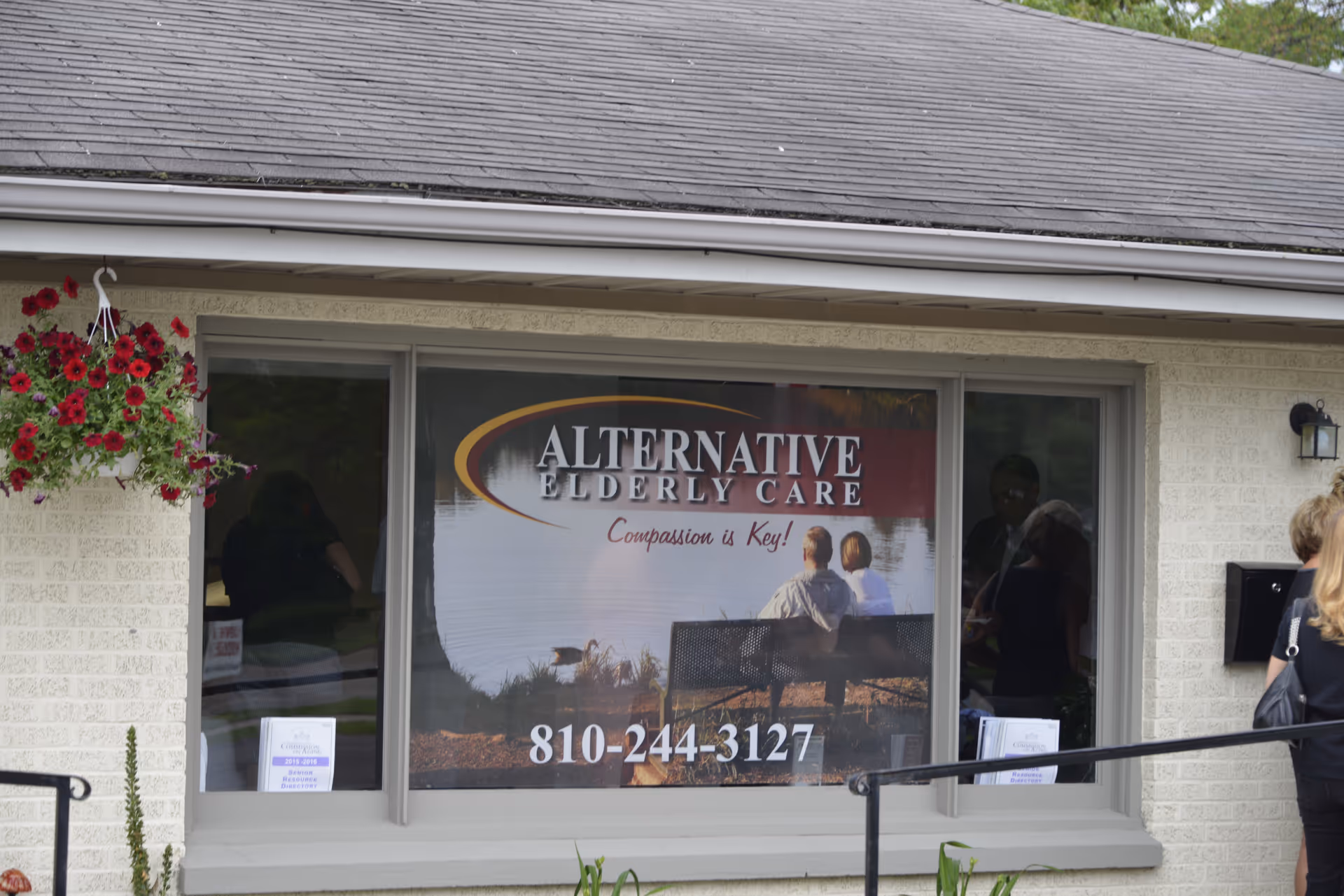 Front view of a building with Alternative Elderly Care window sign and hanging flowers.