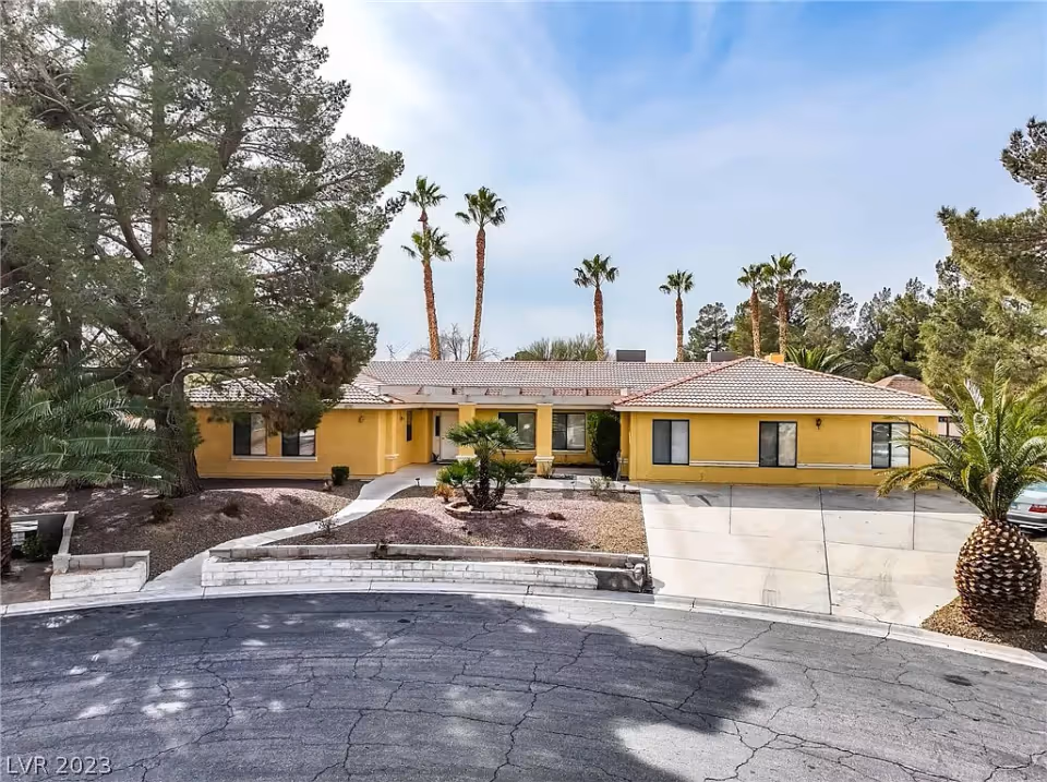 Front exterior view of a single-story yellow building with a tiled roof, surrounded by palm trees and other greenery, with a driveway and a curved road in front.