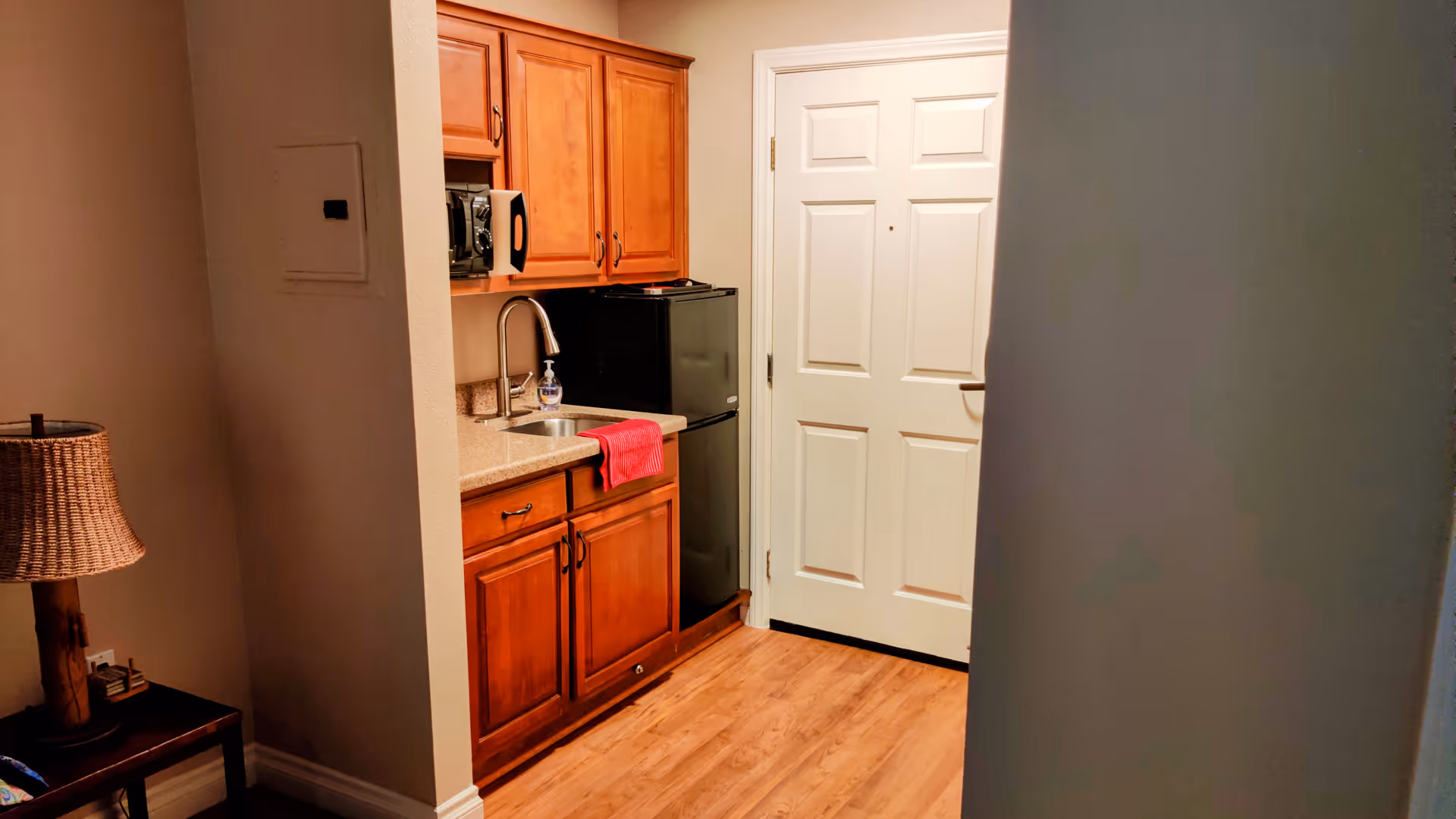 Compact kitchenette featuring wooden cabinets, a sink, microwave, fridge, and a white door.