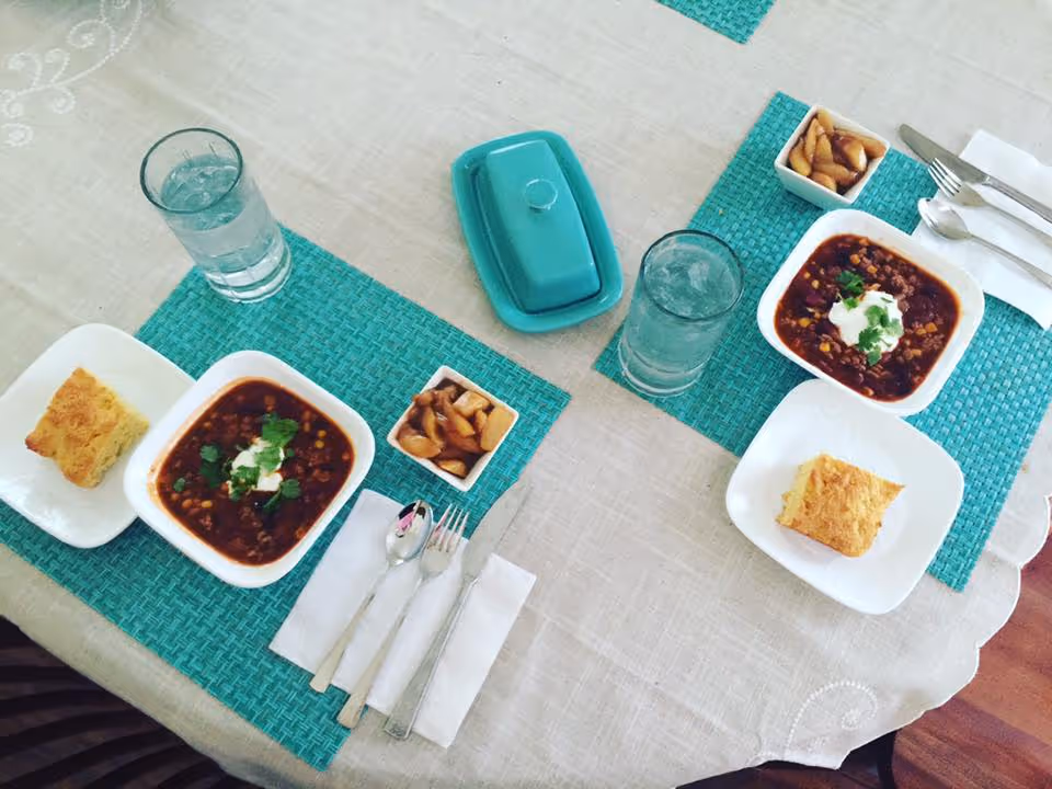 A dining table set for two with turquoise placemats, each setting includes a bowl of chili topped with sour cream and cilantro, a piece of cornbread on a white plate, a small dish of pickled vegetables, a glass of water with ice, and silverware wrapped in a white napkin. A turquoise butter dish is placed in the center of the table.