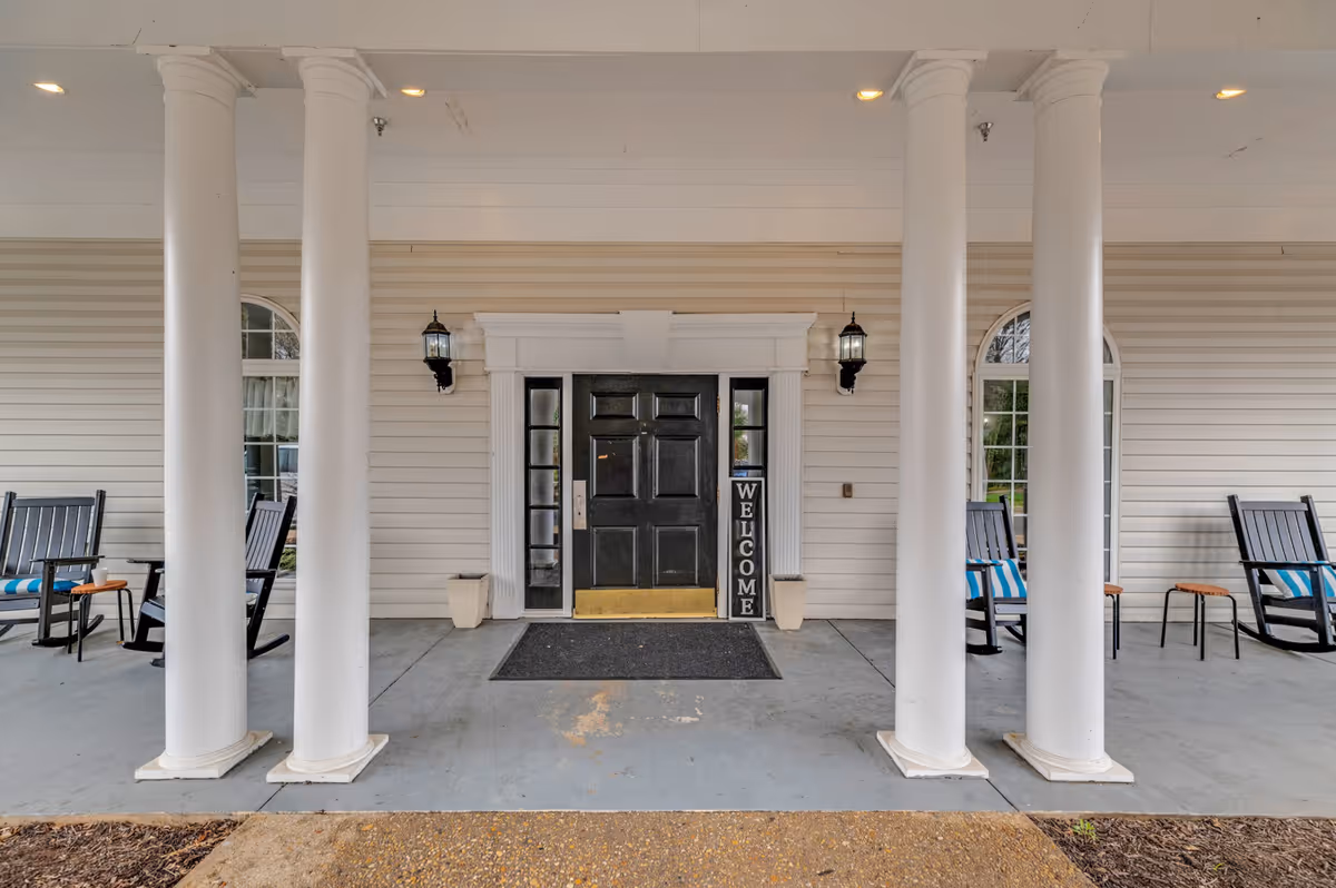 Covered front entrance with white columns, a black door, rocking chairs, and a 'WELCOME' sign.