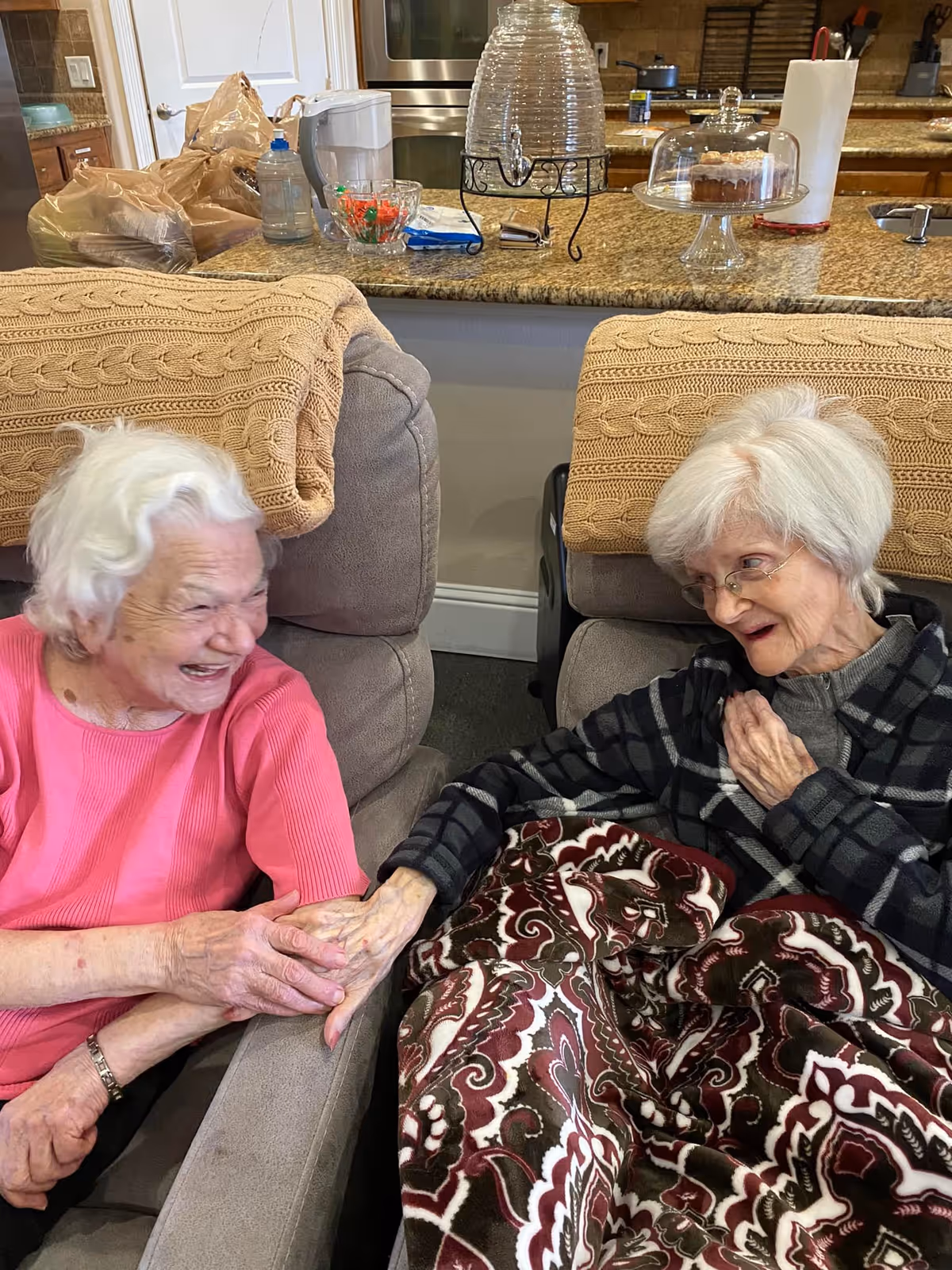 Two elderly women sitting in comfortable armchairs in a living room area, smiling and holding hands. One woman is wearing a pink top and the other is wearing glasses and a dark plaid shirt with a patterned blanket over her lap. Behind them is a kitchen counter with various items including a water dispenser, a cake under a glass dome, and paper towels.